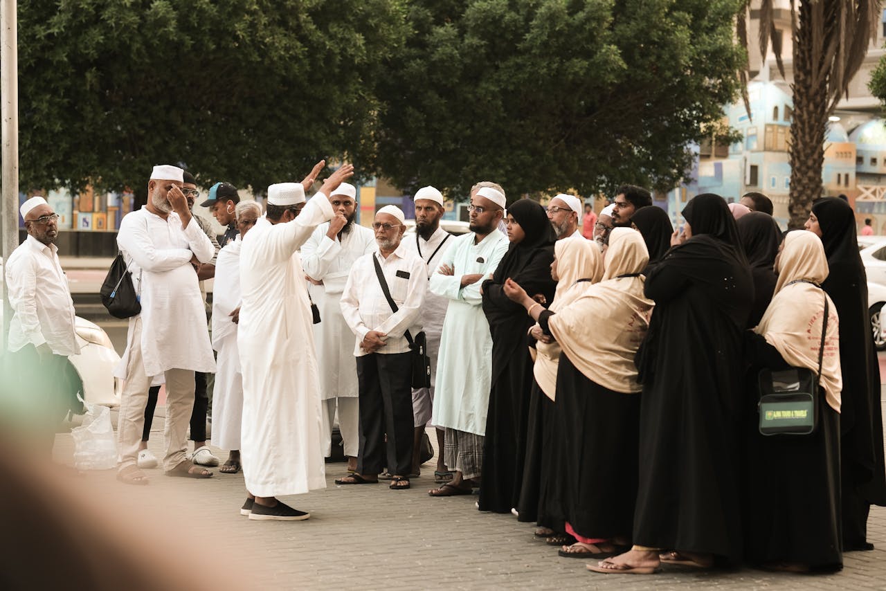 A group of pilgrims in traditional attire gather on a street in Saudi Arabia, engaging in discussion.